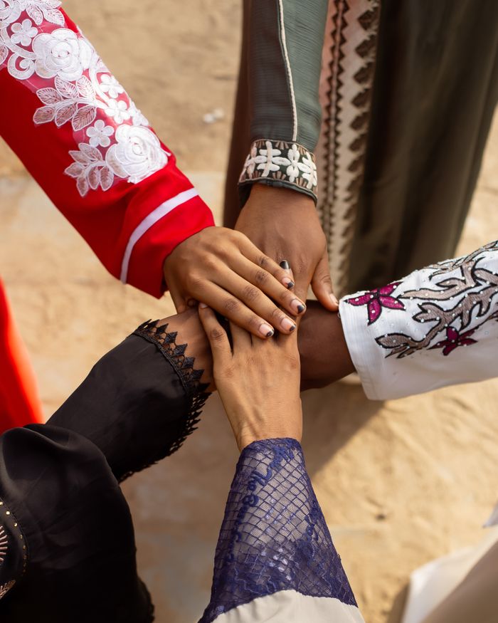 about-01 A group of people joining hands wearing colorful traditional attire outdoors, symbolizing unity.